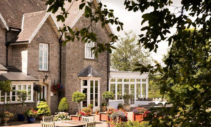 A charming stone house with white-framed windows and a bright conservatory, framed by leafy trees. The patio is filled with colourful flowers, neat to